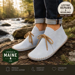 May include: Light blue suede ankle boots with tan leather laces and visible whipstitch detailing along the sole. The boots feature a soft, flexible construction and are worn with dark blue cuffed denim jeans. Text overlays include "LIFETIME REPAIRS & RESOLING! BUILT TO LAST" and "HANDMADE IN MAINE USA".