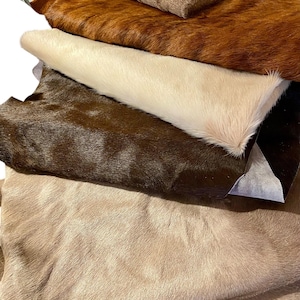 Puede incluir: Una pila de cinco piezas rectangulares de piel de vaca en varios tonos de marr&oacute;n, bronceado, crema y chocolate oscuro. Cada pieza presenta una textura natural de pelo corto, dispuestas una sobre otra para mostrar el degradado de color.