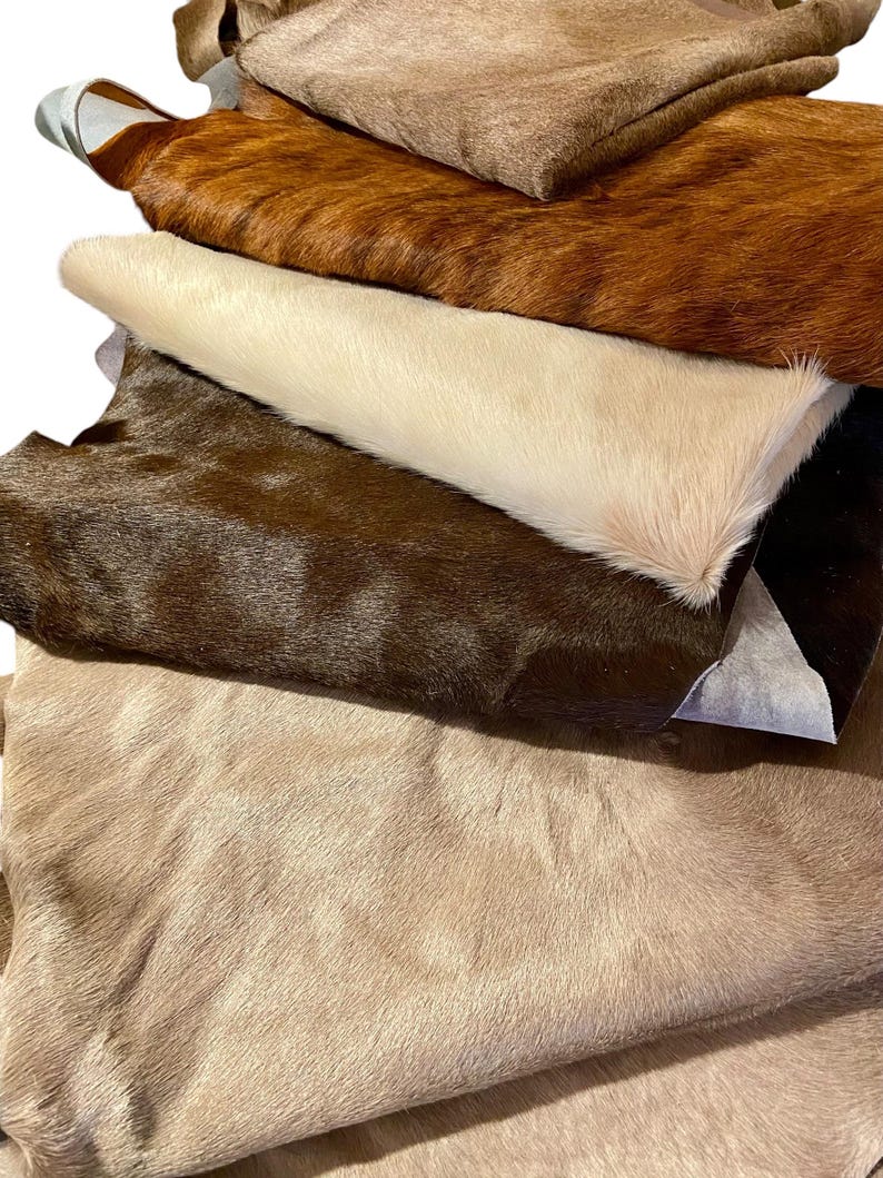 Puede incluir: Una pila de cinco piezas rectangulares de piel de vaca en varios tonos de marr&oacute;n, bronceado, crema y chocolate oscuro. Cada pieza presenta una textura natural de pelo corto, dispuestas una sobre otra para mostrar el degradado de color.