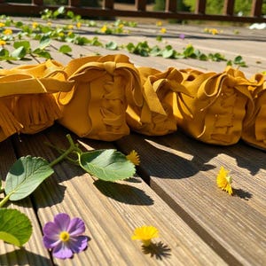 May include: A row of mustard yellow leather moccasins with fringe and decorative stitching. The shoes are arranged on a wooden surface with green leaves, purple and yellow flowers.