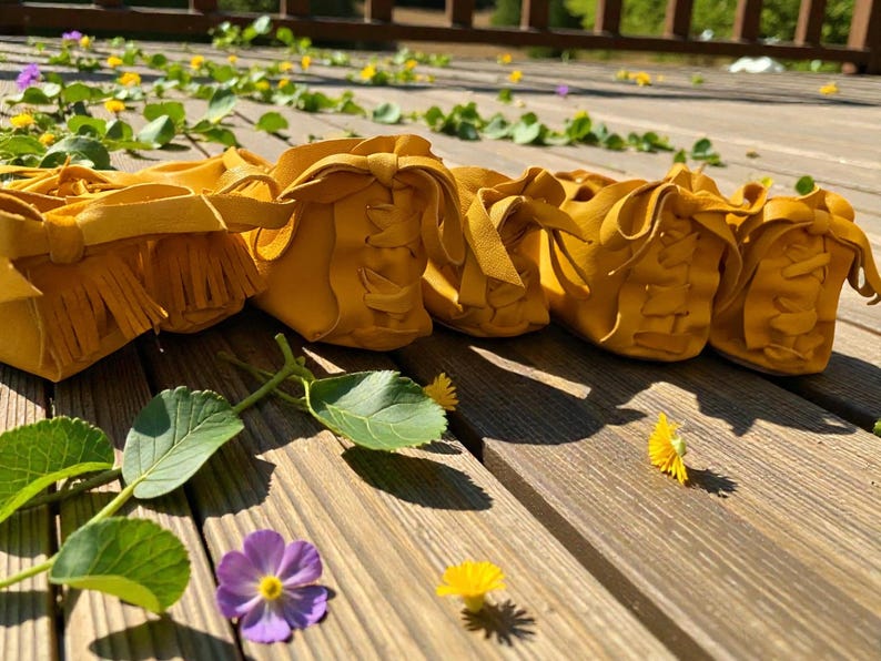 May include: A row of mustard yellow leather moccasins with fringe and decorative stitching. The shoes are arranged on a wooden surface with green leaves, purple and yellow flowers.