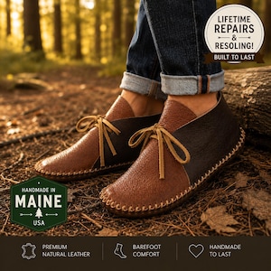 May include: A pair of two-tone brown leather moccasins featuring a dark brown heel and a lighter brown toe section with visible whipstitching along the sole. The shoes have tan leather laces tied in a bow and are worn with cuffed blue jeans.