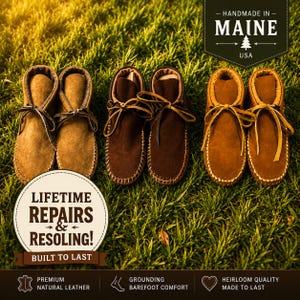 May include: Three pairs of leather moccasin-style shoes in varying shades of brown, arranged on green grass. The shoes feature stitched seams and leather laces. The image includes text that reads "Handmade in Maine, USA" and "Lifetime Repairs & Resoling!"