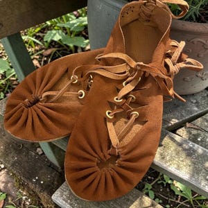 May include: A pair of brown suede moccasin-style shoes with leather laces and brass eyelets. The shoes have a gathered toe design and are displayed on a wooden surface, suggesting a handcrafted or bohemian style.