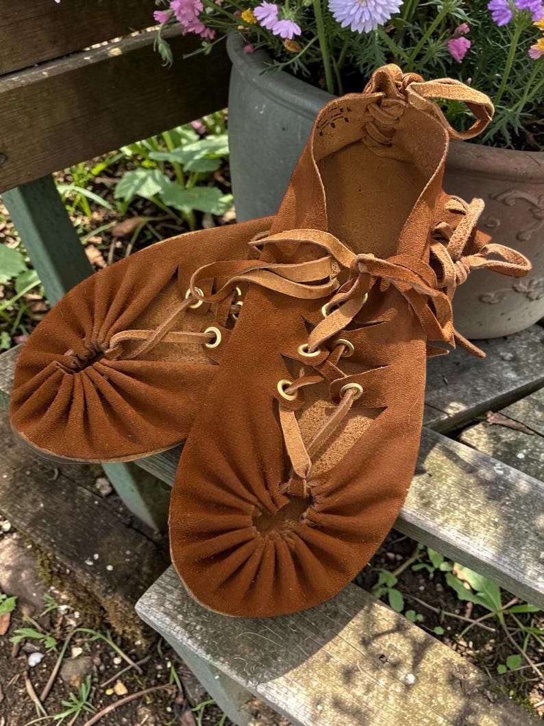 May include: A pair of brown suede moccasin-style shoes with leather laces and brass eyelets. The shoes have a gathered toe design and are displayed on a wooden surface, suggesting a handcrafted or bohemian style.