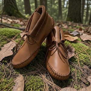 May include: A pair of brown leather moccasin boots with a soft, textured collar and leather laces. The boots are hand-stitched with visible seams and are set against a natural forest backdrop with moss and fallen leaves.