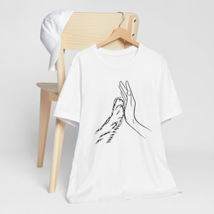 May include: White t-shirt with a black line drawing of a human hand and a paw touching. The shirt is hanging on a wooden hanger. A white baseball cap is on the back of a wooden chair.