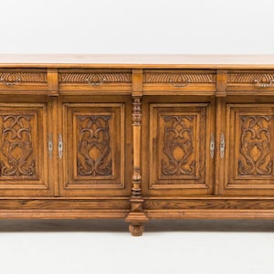 Könnte beinhalten: Ein antikes Sideboard aus Holz mit aufwendigen Schnitzereien. Der Schrank hat vier Türen und zwei Schubladen, alle mit floralen Mustern. Das Holz hat einen warmen, mittelbraunen Ton, und die Beschläge sind silberfarben. Das Sideboard ist etwa 200 cm lang.