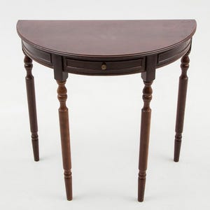 Op de afbeelding: Een donkerbruine, halfronde houten tafel met een lade en vier gedraaide poten. De tafel heeft een klassiek ontwerp, geschikt voor een hal of entree. De tafel is ongeveer 76 cm breed en 76 cm hoog.