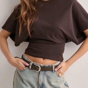 May include: A dark brown, short-sleeved top with a cinched waist. The top is paired with light blue jeans and a brown belt with a silver buckle. The model is wearing gold rings.