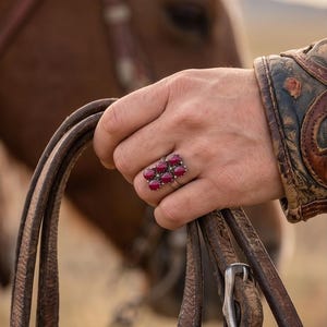 May include: A silver ring with a cluster of oval-shaped, deep red gemstones. The ring is worn on a hand holding leather reins, with a horse and a person's leather-sleeved arm in the background. The ring's gemstones are surrounded by small, clear stones.