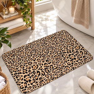 May include: A rectangular bath mat featuring a repeating leopard print pattern with black spots on a tan and beige background. The mat has rounded corners and a flat, low-profile surface, designed for placement on a bathroom floor.