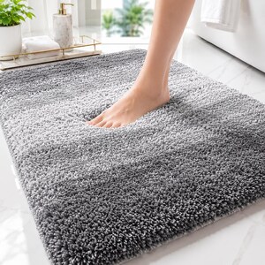 May include: A rectangular bath mat featuring a shaggy, high-pile texture in a gradient of gray tones, transitioning from a lighter shade at the top to a darker shade at the bottom, providing a soft surface for feet to rest upon in a bathroom.