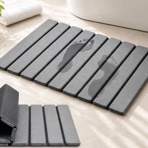 May include: A rectangular gray bath mat composed of multiple parallel slats. The mat features visible wet footprints on its surface. A second, smaller view shows the same gray mat partially folded into a compact, pentagonal roll shape.