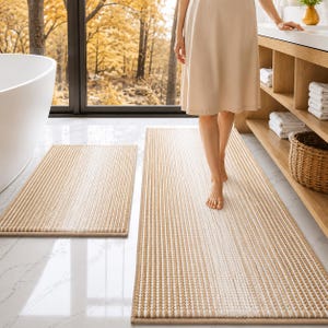 May include: Two rectangular bath mats featuring a textured, ribbed design with alternating vertical stripes of tan and white. The mats have a consistent, uniform weave pattern and are finished with a simple, clean-edged border in a matching tan color.