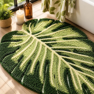May include: A tufted bath mat shaped like a large monstera leaf, featuring a gradient of deep forest green and light lime green fibers. The rug has a textured, shaggy pile surface that mimics the natural veins and split-leaf design of a plant.