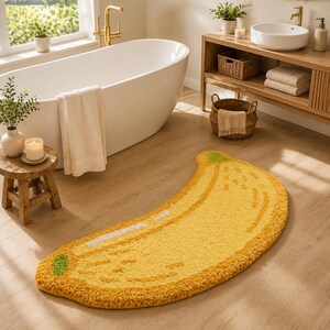 May include: A banana-shaped bath mat featuring a bright yellow tufted texture with a green stem at one end and a white highlight along the curved edge. The rug is designed in the shape of a curved banana, showcasing a soft, plush pile surface.