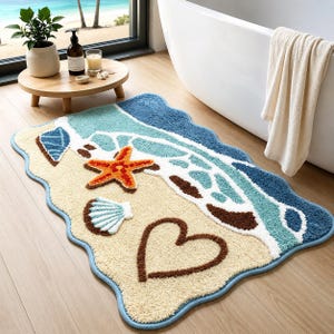 May include: A rectangular bath mat with a wavy edge featuring a beach scene. The design includes a sandy beige area with a brown heart outline, a white seashell, and an orange starfish. The top section depicts blue ocean waves with white foam.