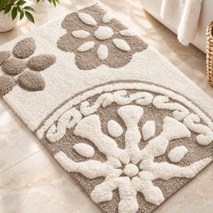 May include: A rectangular bath mat featuring a tufted, high-pile texture in cream and taupe. The design includes a large, semi-circular medallion pattern with radiating petal shapes and two smaller floral motifs in the upper corner.
