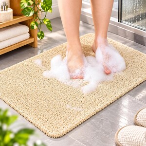 May include: A rectangular beige bath mat with a textured, porous surface design. The mat features a loofah-like construction that allows water to drain through. It is positioned on a tiled floor, providing a non-slip surface for feet.