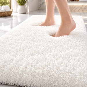 May include: A rectangular white bath mat with a thick, high-pile shaggy texture. The mat features a dense, soft surface that conforms slightly under pressure, showcasing its plush construction and deep, absorbent fibers for use in a bathroom.