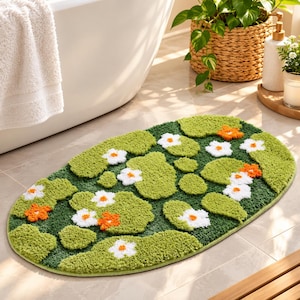 May include: An oval-shaped bath mat featuring a tufted texture with a dark green background. The design includes light green lily pad shapes interspersed with small white flowers and orange flowers scattered across the surface of the mat.