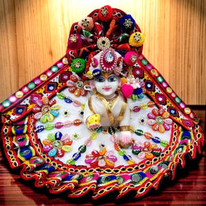 May include: A detailed, ornate doll dressed in a white and gold outfit, adorned with colorful embroidery, mirrors, and floral accents. The doll wears a decorative headpiece with jewels and pom-poms. The overall design is vibrant and festive.