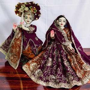 May include: Two ornate figurines dressed in deep purple and copper-toned fabric with intricate gold embroidery and sequins. The left figure wears a large, embellished headpiece and holds a flute, while the right figure wears a draped head covering.