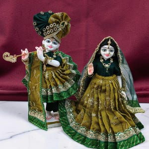 May include: Two ornate figurines of a couple, possibly religious figures, are displayed. The figures are dressed in elaborate green and gold clothing with intricate embroidery and embellishments. One figure holds a flute. The background is a deep red.