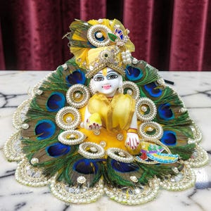 May include: A small, white figurine of a deity wearing a yellow outfit and a jeweled crown. The figurine is adorned with peacock feathers and pearl accents. The background is a marble surface and a burgundy curtain.