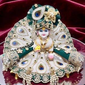 May include: A white marble figurine of a deity dressed in an elaborate green and white outfit. The attire features peacock feather motifs, gold embroidery, pearl accents, and a matching turban with a large decorative peacock feather embellishment.