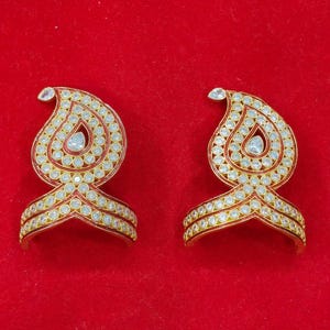 May include: A pair of gold-toned earrings featuring a teardrop-shaped paisley design. Each earring is embellished with numerous small, clear, round-cut stones and a single larger teardrop-shaped stone in the center, set against a red enamel base.