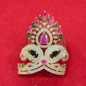 May include: A gold-toned ring featuring a large, ornate, crown-like design encrusted with clear white stones. The piece is accented with pear-shaped pink and dark green stones arranged in a symmetrical pattern, creating a bold, decorative statement.