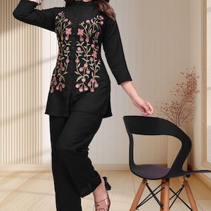 May include: A black long-sleeved tunic featuring a button-down front and intricate floral embroidery in shades of pink and green, paired with matching black trousers. The tunic has a high collar and a straight hemline that reaches the mid-thigh.