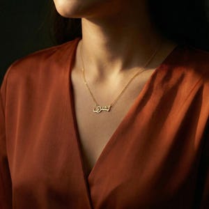 Pode incluir: Um colar dourado com a palavra árabe "بشرى" (Bushra) como pingente. O colar é usado sobre uma blusa cor de ferrugem. A corrente é delicada e o pingente está em fonte cursiva.