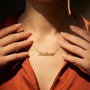 以下が含まれることがあります： 筆記体で「Melissa」と書かれたゴールドカラーのネックレス。ネックレスは人の首に着用され、手でそっと触れています。その人はオレンジがかった赤色のトップスを着ています。