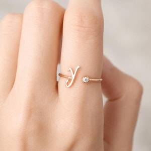 Puede incluir: Un anillo abierto de oro rosa que presenta una letra Y en cursiva en un extremo y una pequeña piedra preciosa redonda y transparente engastada en bisel en el otro. La banda es fina y pulida, creando un aspecto delicado en el dedo.