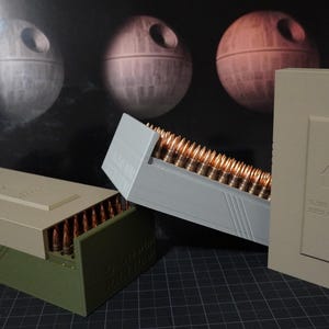May include: Three ammunition storage containers in shades of green, gray, and tan. The containers are rectangular and hold numerous copper-colored bullets. The background features a dark image of three Death Star-like spheres.