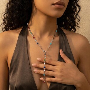 Può includere: Una collana color argento con una serie di pietre preziose colorate. La collana presenta un design a forma di Y, con una fila di gemme a cascata al centro. Le pietre preziose sono di vari colori, tra cui rosso, blu, verde e giallo.