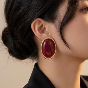 May include: An oval-shaped earring featuring a smooth, deep red cabochon stone set within a polished gold-toned metal frame. The earring has a classic, rounded silhouette and is worn on the earlobe, showcasing a glossy finish and rich colour.