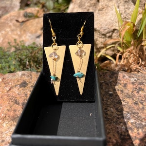 Peut inclure: Une paire de boucles d'oreilles dorées de forme triangulaire. Chaque boucle d'oreille est ornée d'un cristal transparent, d'une petite perle turquoise et d'une chaîne dorée. Les boucles sont présentées dans un coffret noir, sur fond naturel.