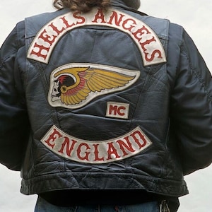 May include: Black leather jacket with embroidered patches. The patches feature the words "HELLS ANGELS" in red lettering, a winged skull emblem, and "ENGLAND". The jacket also has an "MC" patch.