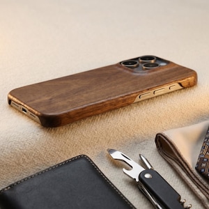 May include: A smartphone case crafted from dark brown wood with a visible grain pattern, featuring a cutout for a triple-lens camera system and metallic side buttons, positioned at an angle to display the back and side profile of the device.