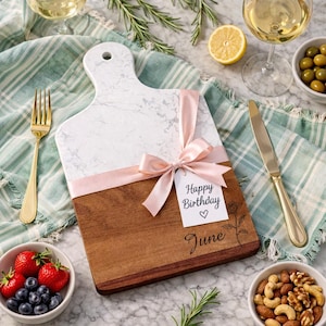 Puede incluir: Una tabla de cortar de dos tonos de madera y mármol con una cinta rosa y una etiqueta que dice "Happy Birthday June". La tabla está rodeada de comida y bebida, incluyendo fruta, nueces, aceitunas y vino.