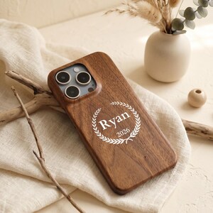 Puede incluir: Una funda de madera de color marrón oscuro para teléfono con un diseño de corona de laurel grabado en láser blanco que rodea el nombre Ryan y el año 2026. La funda tiene un recorte para un sistema de triple cámara en la esquina superior izquierda.