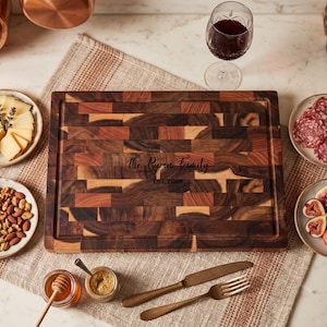 Puede incluir: Una tabla de cortar de madera con un patrón de mosaico marrón oscuro y marrón claro. El texto "The Ryan Family Est. 2026" está grabado en la tabla. La tabla está rodeada de platos de queso, nueces, salami, fruta, miel y una copa de vino tinto.