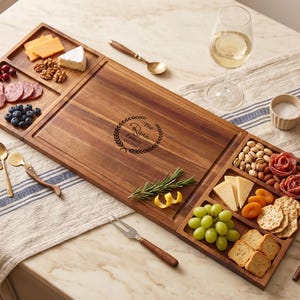 Puede incluir: Una tabla de servir de madera con compartimentos llenos de quesos, carnes, nueces, frutas y galletas. La tabla tiene un grabado personalizado con el nombre de la familia. También hay cucharas doradas y una copa de vino.