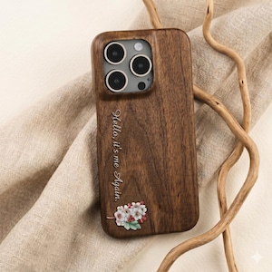 Puede incluir: Funda de teléfono de madera marrón oscuro con diseño floral. La funda tiene el texto grabado "Hello, it's me Again." y una pequeña ilustración floral con flores blancas y bayas rojas.