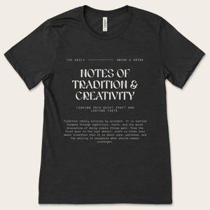 May include: Dark grey t-shirt featuring the white text "NOTES OF TRADITION & CREATIVITY". The shirt also includes the phrases "THE DAILY" and "SMIRK & DRINK". The design is a statement about craft and taste, with a focus on tradition.