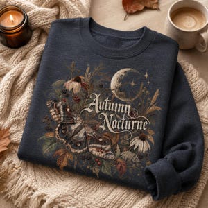 May include: A dark navy blue crewneck sweatshirt featuring a graphic print of a large moth, a crescent moon, and various autumn wildflowers. The text "Autumn Nocturne" is printed in a decorative script font across the center of the design.